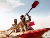 Young couple sharing a red kayak smiling and laughing while paddling through shallow ocean water in New Jersey