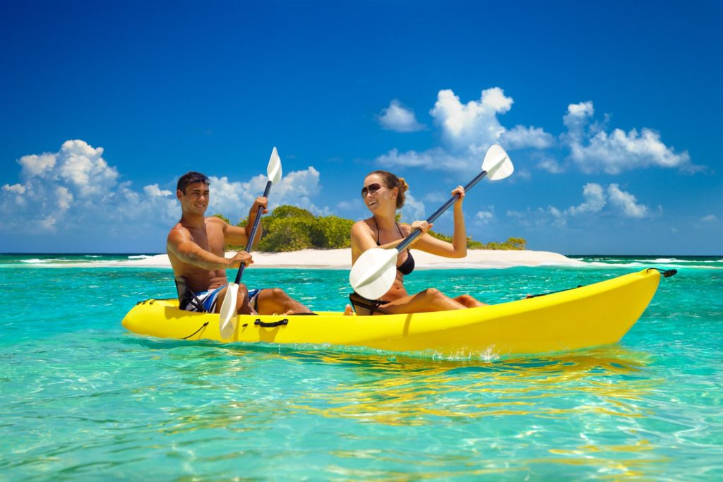 A young couple sharing a yellow kayak and paddling through shallow ocean water past a small island