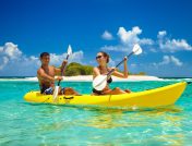 A young couple sharing a yellow kayak and paddling through shallow ocean water past a small island