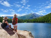 Two women standing on a rock overlooking a large blue lake, a dense green forest and mountain peaks on a sunny day