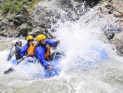 A group of kayakers with jackets, helmets, and life vests paddling a large blue kayak through whitewater rapids