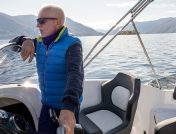 Older man in a blue jacket driving a boat over a calm lake during winter