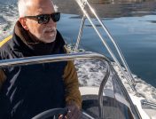 Older man in a heavy coat driving a boat over a calm lake during winter